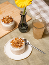 Two small oat dishes with granola, a glass of iced tea, and a vase with a yellow flower on a textured surface.