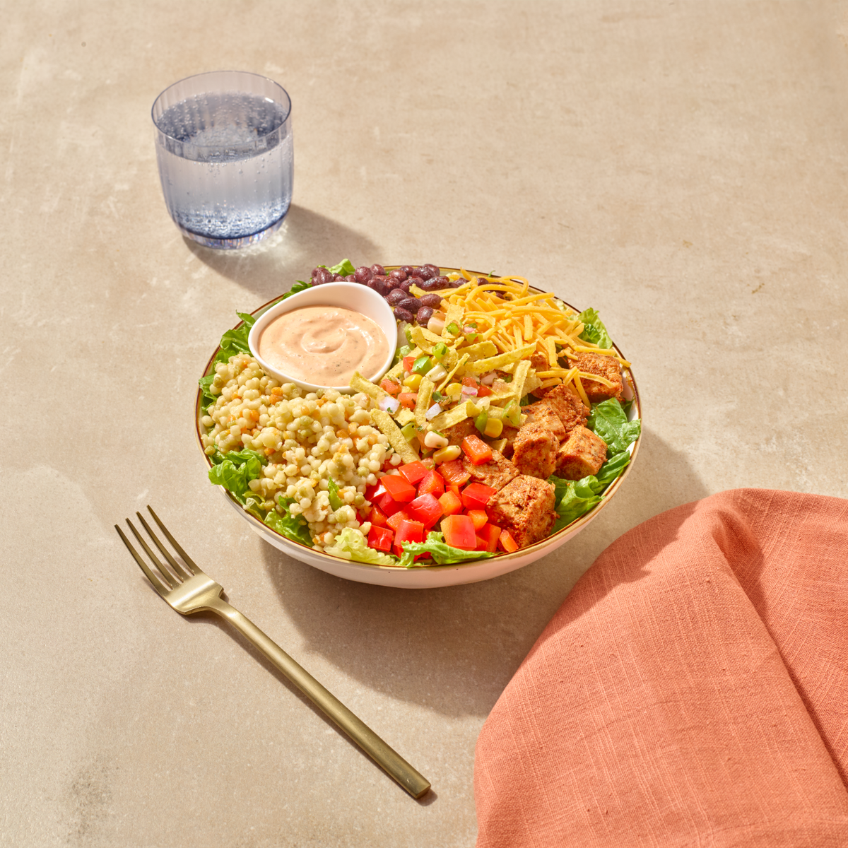 A colorful salad bowl filled with mixed greens, corn, diced tomatoes, shredded cheese, and chicken, served on a beige stone-like surface with a gold fork, a blue glass, and a peach napkin.
