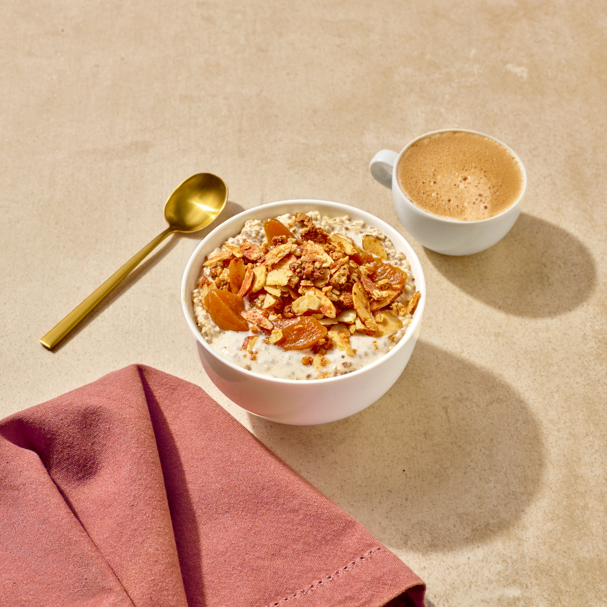 A styled breakfast scene on a beige surface featuring a white bowl of oatmeal topped with nuts and dried fruit, a white cup of coffee, a gold spoon, and a folded dusty rose napkin.