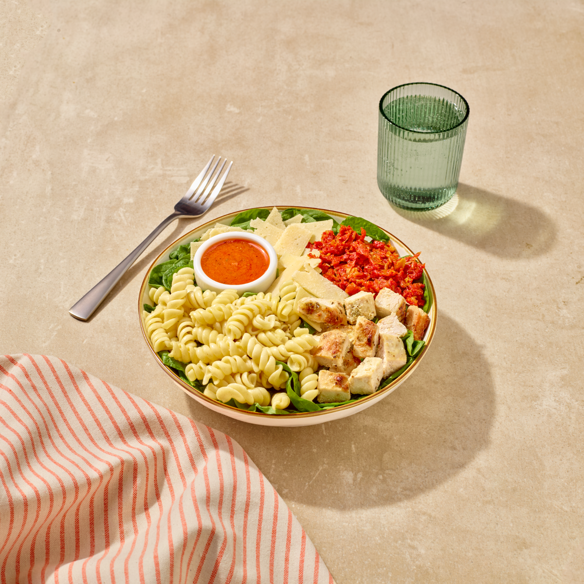A prepared pasta salad bowl with mixed greens, rotini pasta, chicken, red peppers, and a small orange dipping sauce, styled on a beige surface with a fork, glass, and striped napkin.