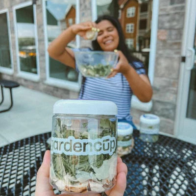 Close-up of a GardenCup™ salad jar on an outdoor table, with a smiling woman enjoying her fresh salad in the background. Showcases the freshness and convenience of GardenCup’s healthy salad delivery service.